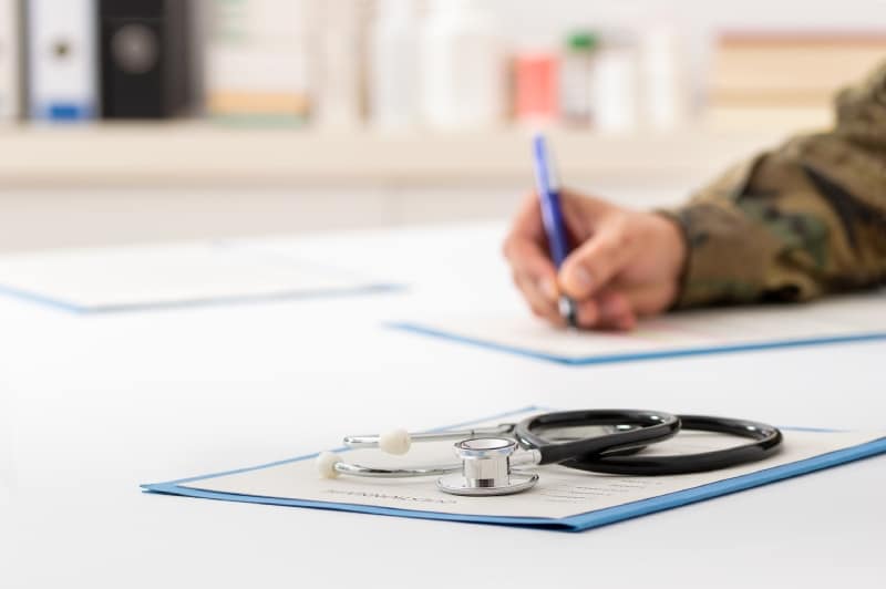 Cropped picture army person completing a form in background with stethoscope and paperwork on white table