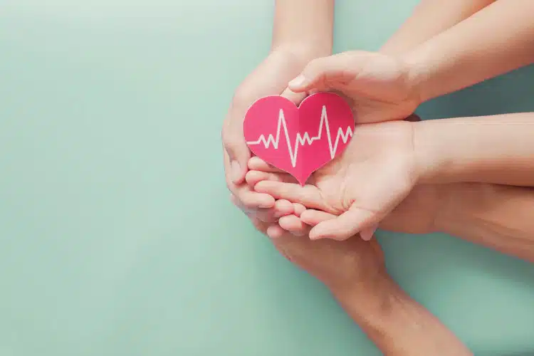 Tres juegos de manos de adultos y niños sosteniendo un corazón rojo con una línea blanca de frecuencia cardíaca a lo largo
