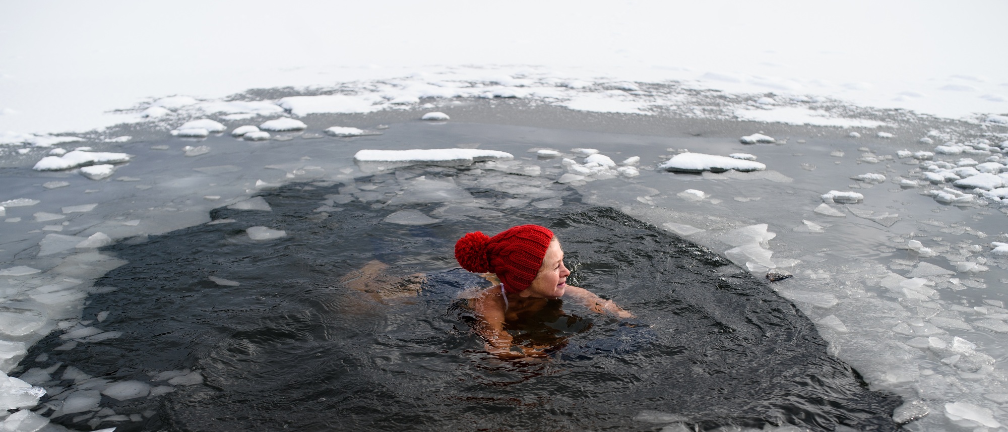 Active senior woman swimming in water hole in frozen lake outdoors in winter, cold therapy concept.