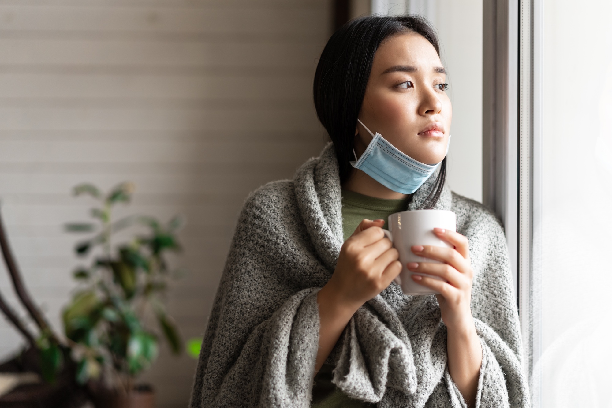 Sick asian girl in medical face mask standing by the window and yearning to go outside, being on